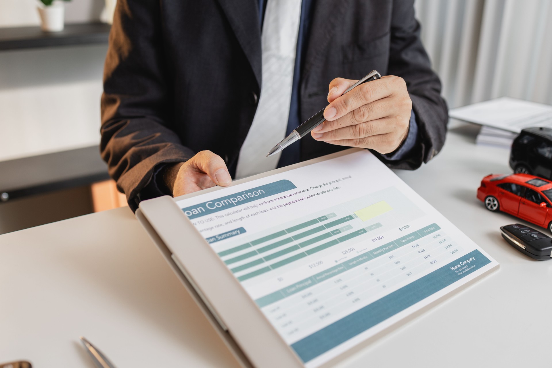 A businessman in a suit is calculating loan details, reviewing financial documents, and holding car keys near a calculator and toy car models, representing auto loan and finance services.